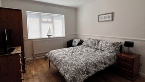 Ein Schlafzimmer mit Bett, Schminktisch, Nachttisch, Fenster mit Jalousien, Heizkörper und Deckenleuchte.