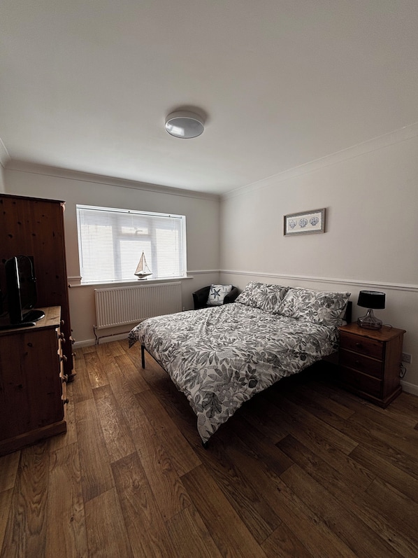 A bedroom with a bed, a dresser, a nightstand, a window with blinds, a radiator, and a ceiling light fixture.