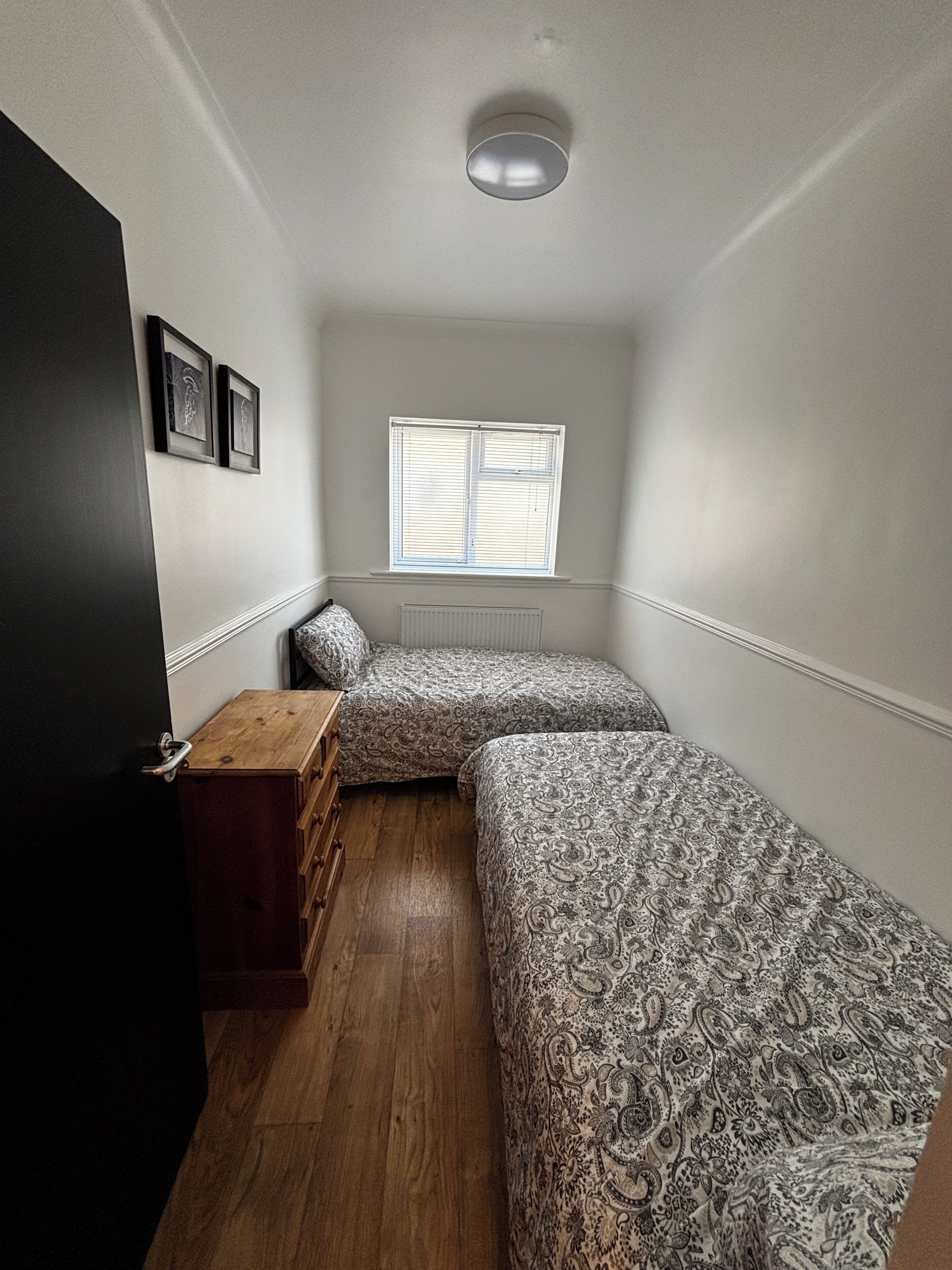 A bedroom with two beds, a wooden dresser, a radiator, and a window with blinds.