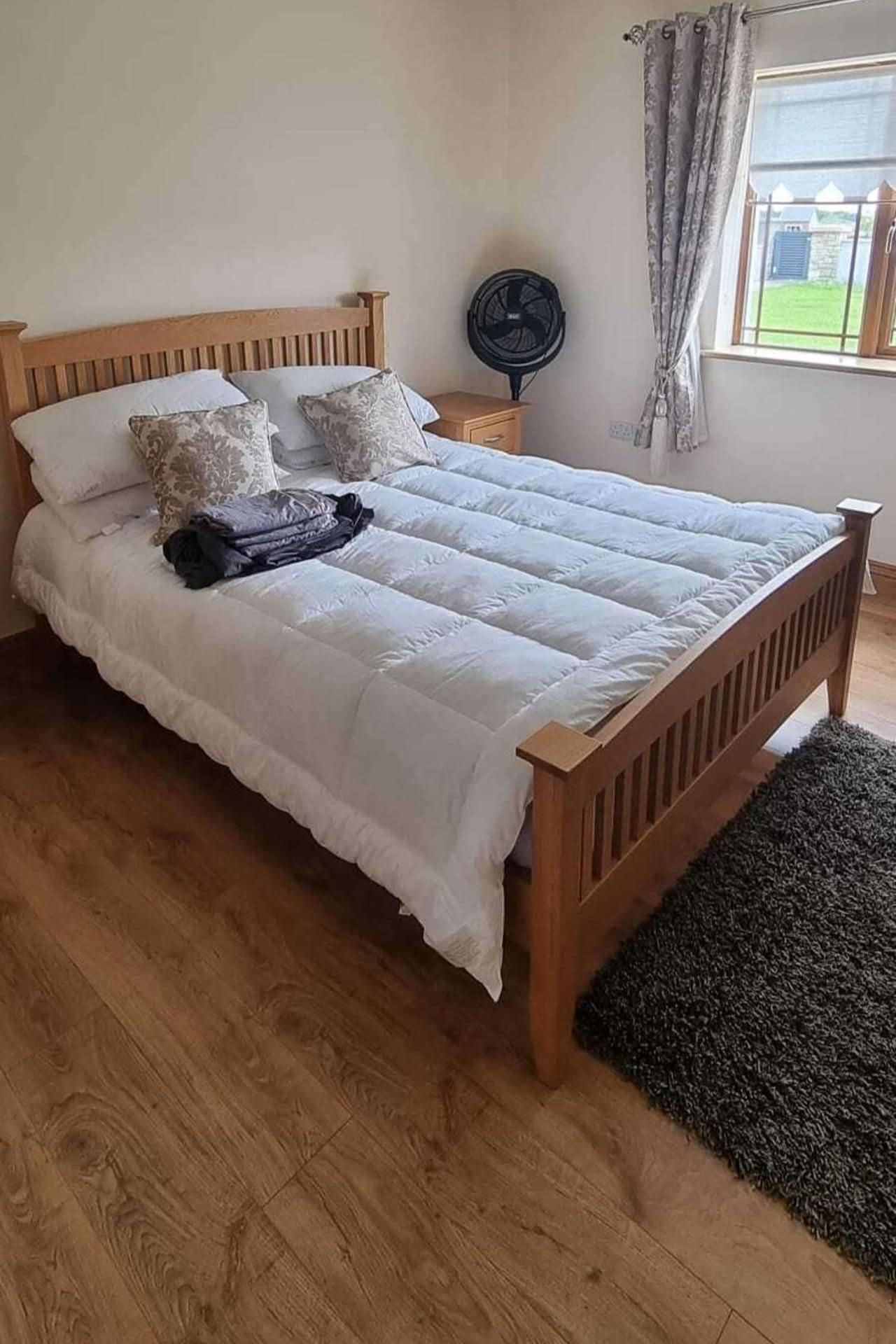 A bed with a wooden frame and white bedding, a small bedside table with a fan, and a window with curtains.