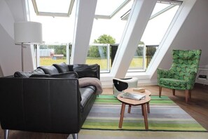 En moderne stue med en sort lædersofa, en grøn stilet stol, et rundt sofabord og udsigt gennem ovenlys.