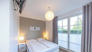 Ett sovrum med en säng, sängbord, en väggmonterad lampa, ett fönster med gardiner och utsikt över grönområden utanför.