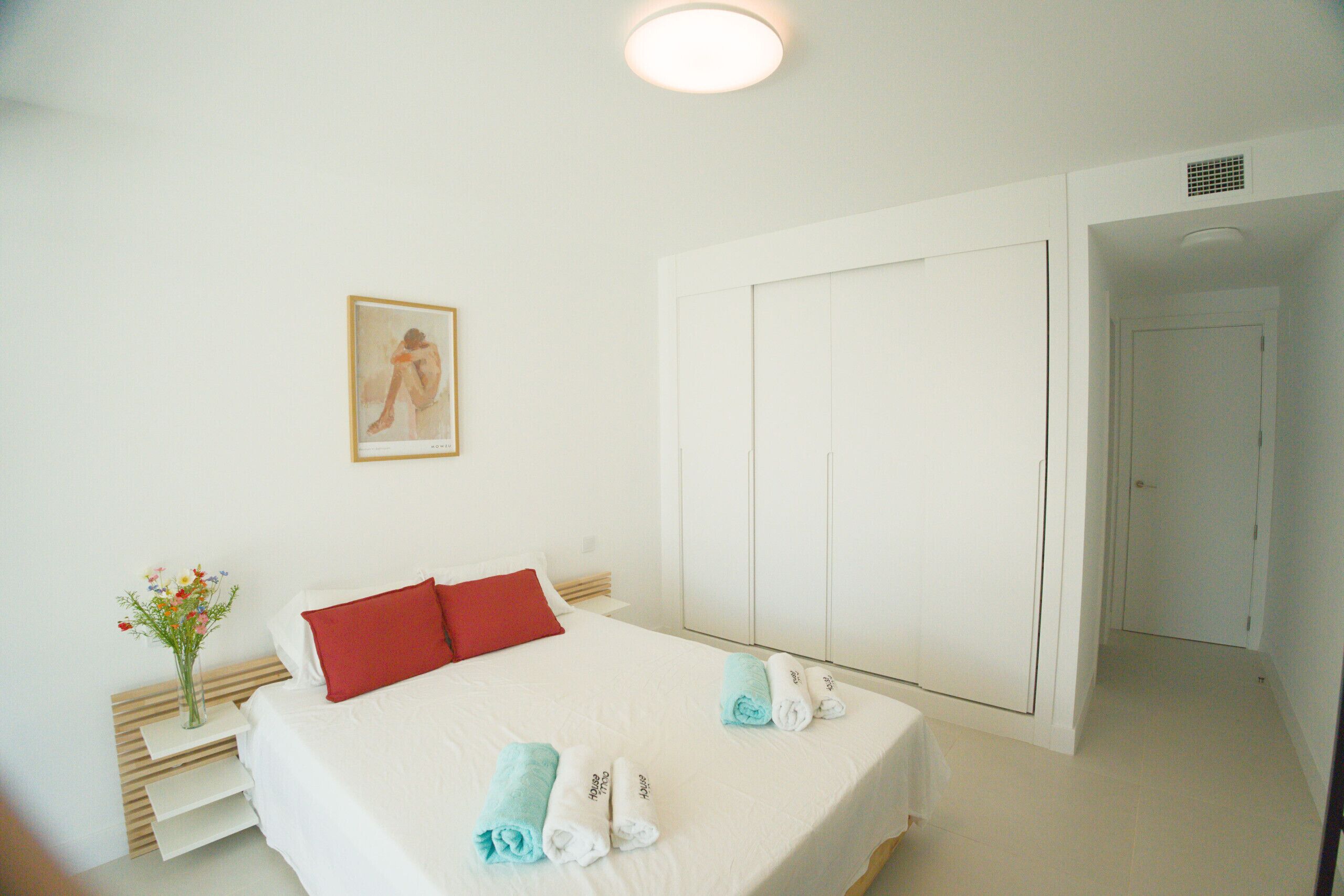 Een hotelkamer met een bed, een nachtkastje met een bloevas, een rode kussen en een witte kast.