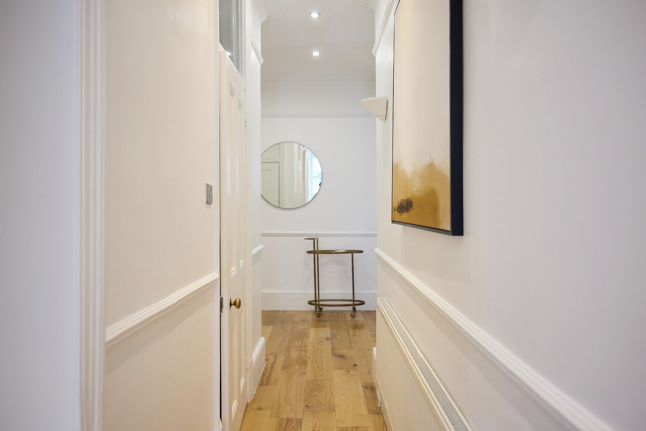 A hallway with a wooden floor, a round mirror on the wall, and a metal rack.
