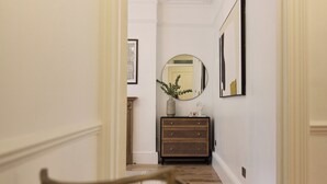 A hallway with a wooden floor, a mirror on the wall, a small wooden cabinet, and a chair in the foreground. - Sage & Ivory - Maida Vale (London)