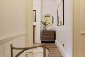 A hallway with a wooden floor, a mirror on the wall, a small wooden cabinet, and a chair in the foreground. - Sage & Ivory - Maida Vale (London)