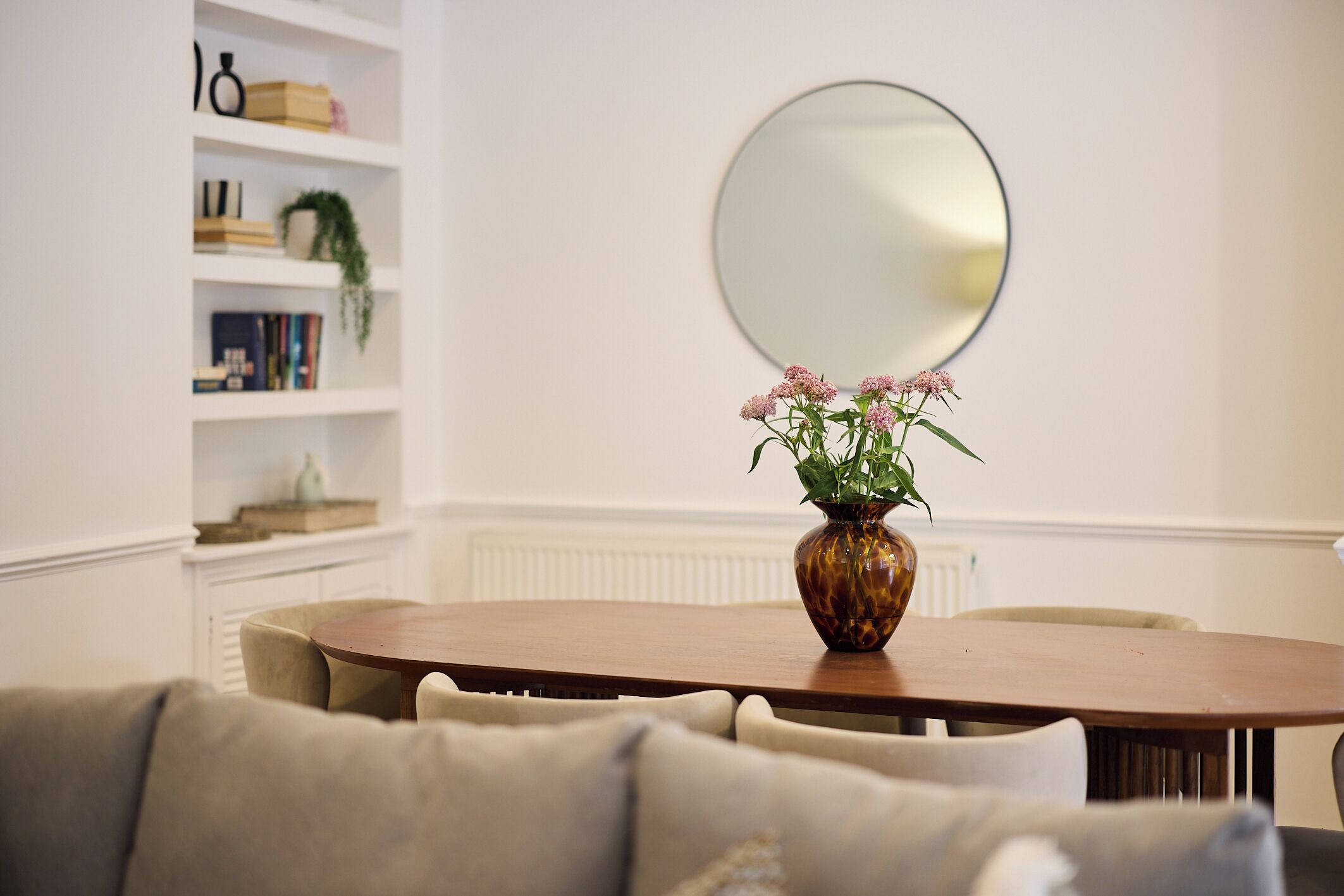 A dining area with a round table, chairs, a vase with flowers, a bookshelf, and a mirror on the wall.