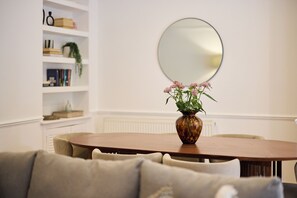 A dining area with a round table, chairs, a vase with flowers, a bookshelf, and a mirror on the wall. - Sage & Ivory - Maida Vale (London)