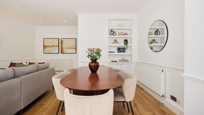 A dining area with a round table, white chairs, a vase with flowers, a sofa, and a bookshelf. - Sage & Ivory - Maida Vale (London)