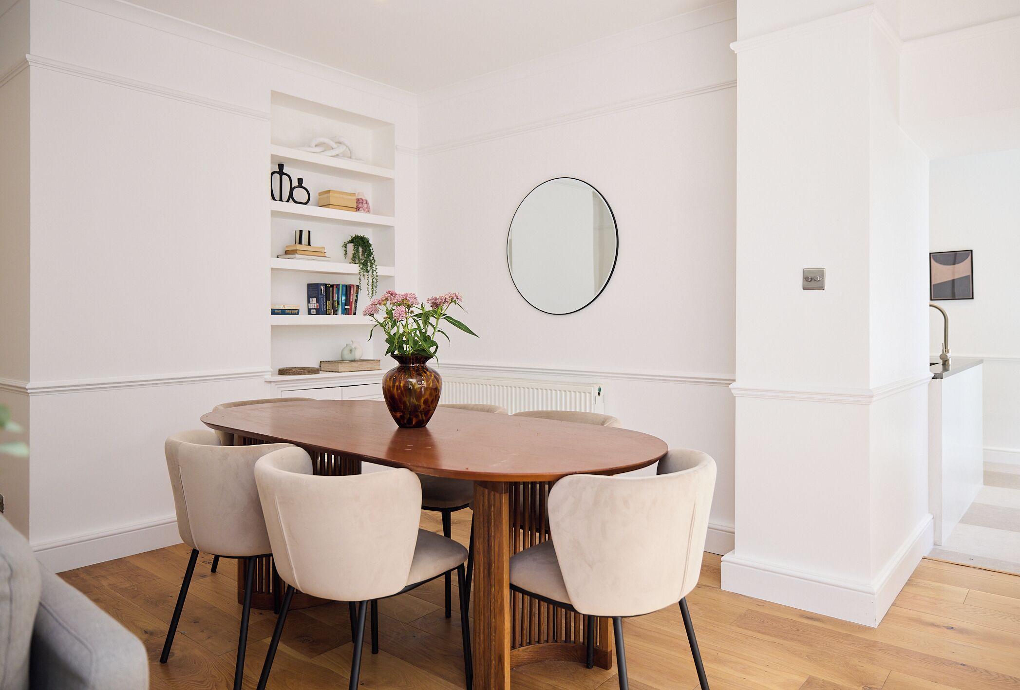 A dining area with a wooden table, four chairs, a vase with flowers, and a mirror on the wall.