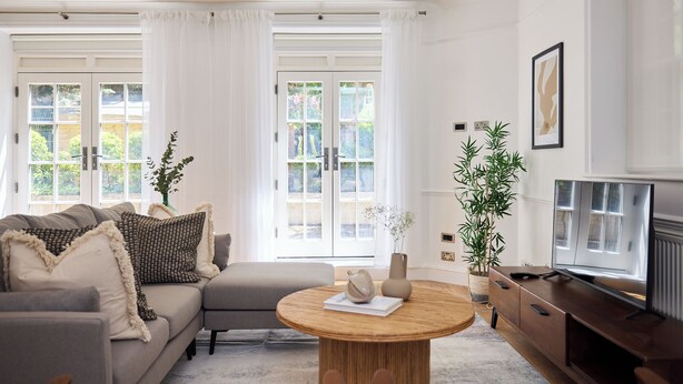 A living room with a sofa, a round coffee table, a TV, and large windows with curtains. - Sage & Ivory - Maida Vale (London)