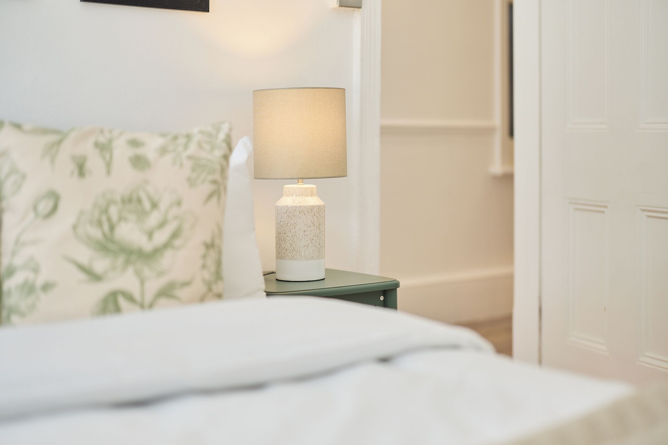 En seng med hvid dyne, et grønt natbord med en lampe, og en pude med blomstermønster.