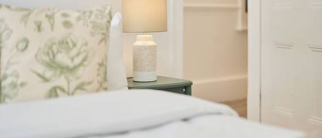 En seng med hvid dyne, et grønt natbord med en lampe, og en pude med blomstermønster.