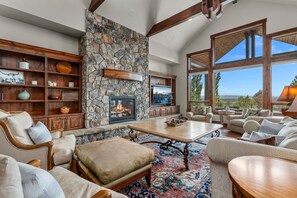 A spacious living room with a stone fireplace, wooden shelves, comfortable seating, and large windows offering a view.