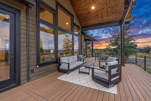 A covered outdoor deck with a sofa and coffee table, large windows, and a view of the sunset.