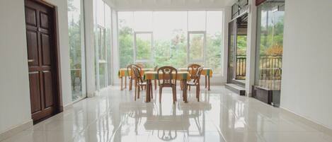 Una habitación amplia con suelo de baldosas, una mesa de comedor con sillas y ventanas grandes que ofrecen vistas a zonas verdes.