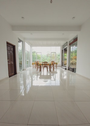 A spacious room with a tiled floor, a dining table with chairs, and large windows offering a view of greenery. - Nilaro Holiday Resort (Anuradhapura)