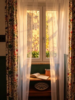 A room with a window covered by sheer curtains, a bowl, and an open book on a wooden surface.