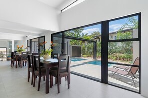 A modern dining area with a large table and chairs, a vase with flowers, and a view of a swimming pool and garden through large glass doors. - Side by Side Villas Walking Distance to Both Playa Tamarindo & Playa Langosta (Playa Langosta)