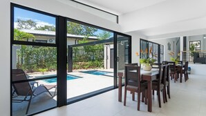 A modern dining area with a pool view, large sliding glass doors, and a kitchen in the background. - Side by Side Villas Walking Distance to Both Playa Tamarindo & Playa Langosta (Playa Langosta)