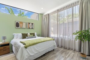 Ein Schlafzimmer mit Bett, Nachttisch, Lampe, Fenster mit Vorhängen und einer Pflanze.