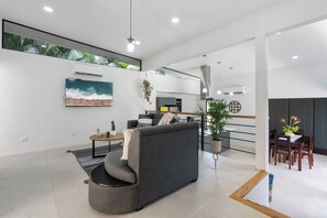Ein modernes Wohnzimmer mit einem grauen Sofa, einem Couchtisch und einem an der Wand befestigten Fernseher. Es gibt einen Essbereich mit einem Holztisch und Stühlen.