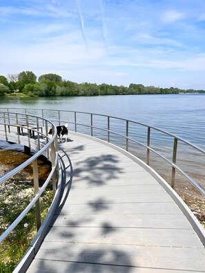 Ein Holzsteg entlang eines Flusses mit Metallgeländern, umgeben von viel Grün.