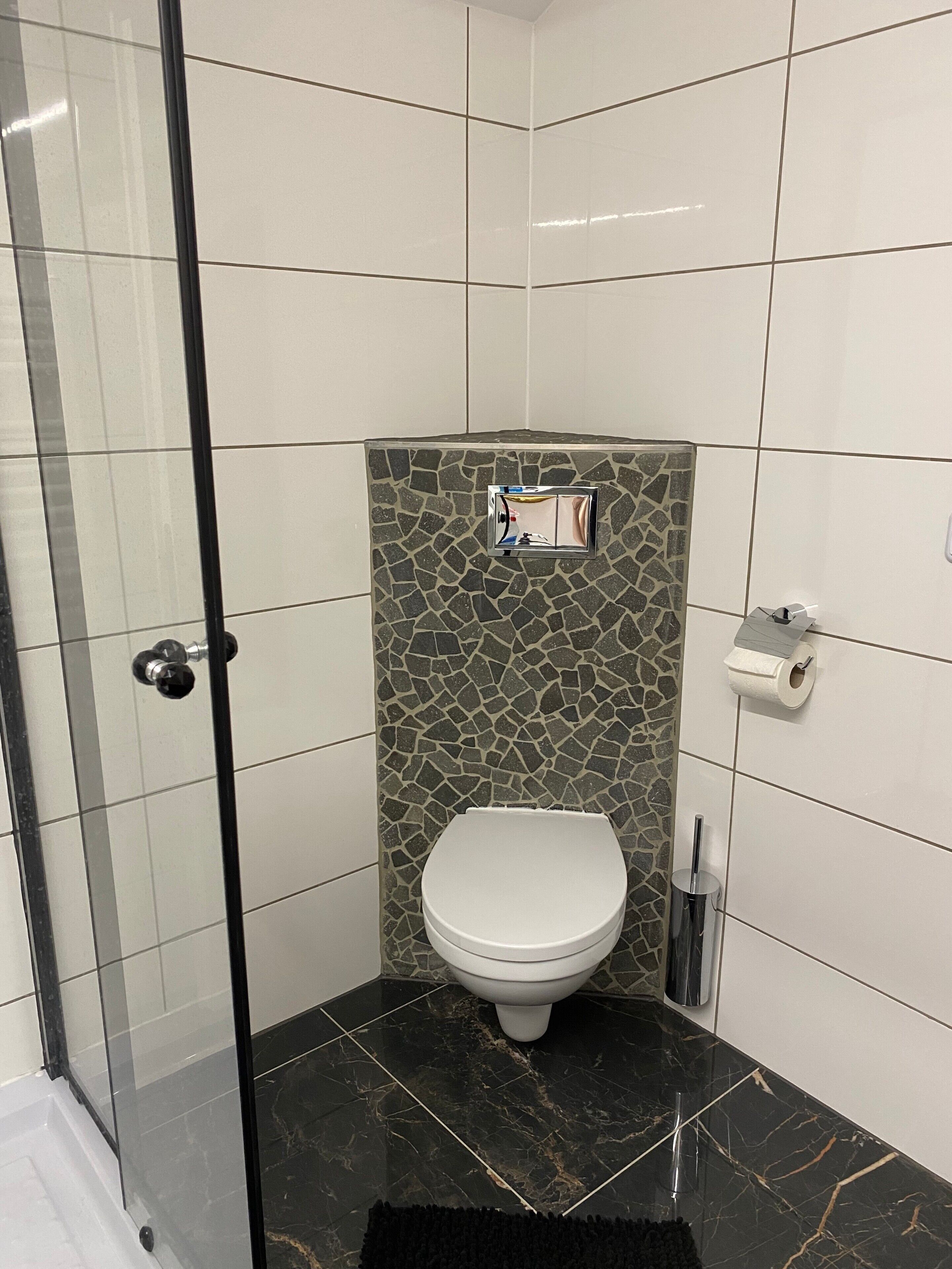 Une salle de bain avec des toilettes blanches, une douche dotée d’une porte en verre et un mur recouvert de mosaïques.