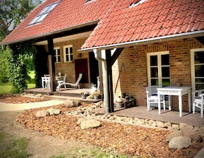 Une maison en briques avec un toit recouvert de tuiles rouges, un porche avec des chaises blanches et un chemin en pierre.