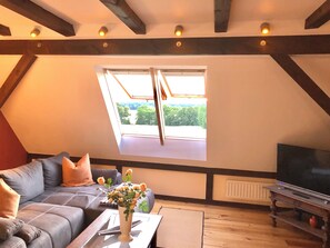 Une pièce de style loft avec un plafond en pente, une grande fenêtre donnant sur un espace verdoyant, un canapé avec des coussins, un vase rempli de fleurs et un téléviseur posé sur un meuble en bois.