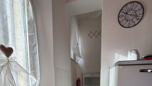 A kitchen with white tiled walls, a white refrigerator, a clock, and a window with sheer curtains.