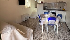 A compact room with a bed, a dining table with chairs, a kitchenette, a TV mounted on the wall, and a staircase leading to an upper level.