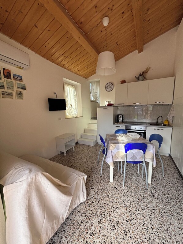 Une pièce de taille modeste comprenant un lit, une table à manger avec des chaises, un coin cuisine, un téléviseur fixé au mur et un escalier menant à un niveau supérieur.
