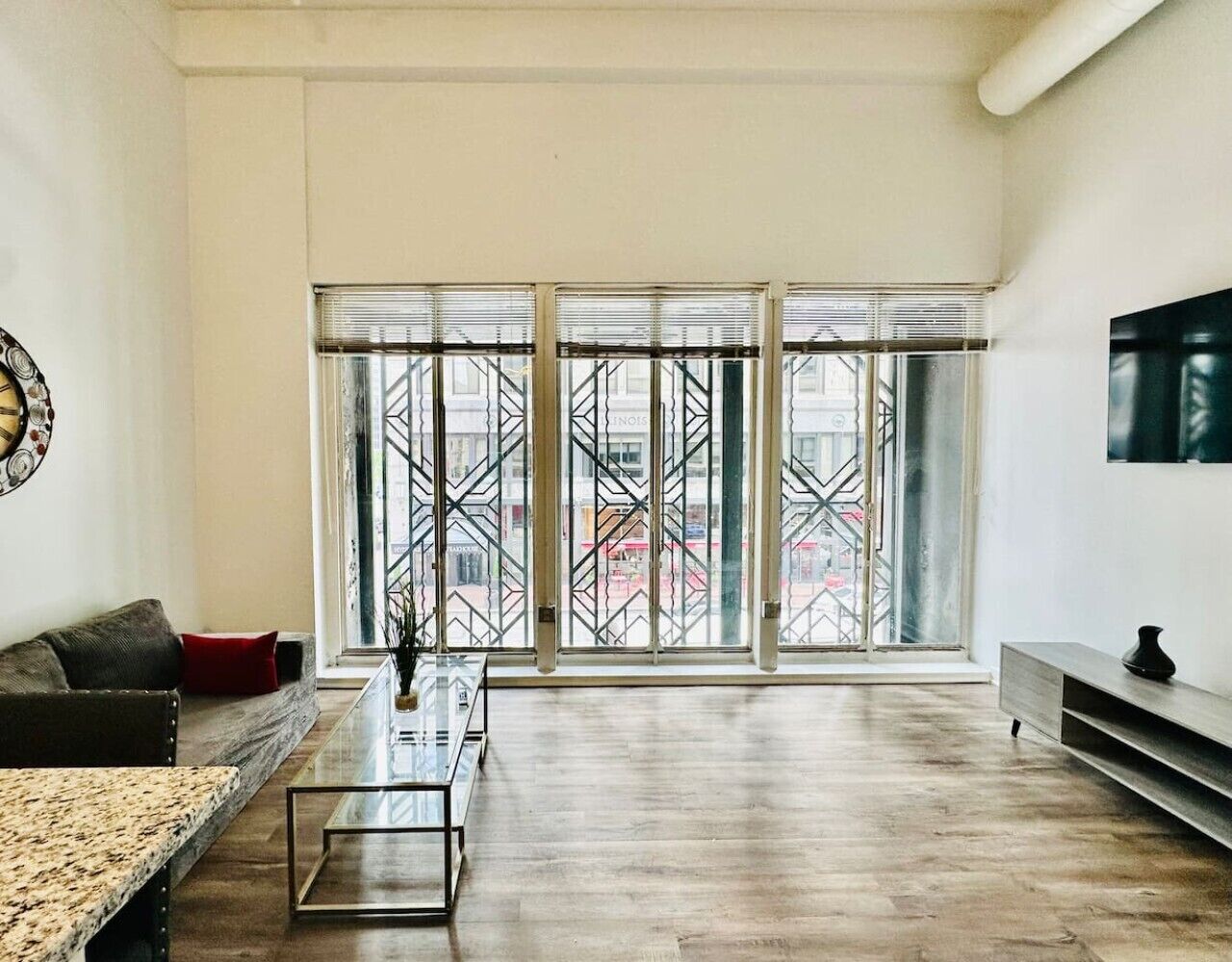 A spacious living room with a large window, a sofa, a coffee table, and a TV mounted on the wall.