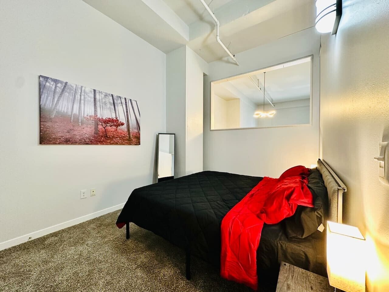 A bedroom with a bed, a red blanket, a painting, a mirror, and a light fixture.