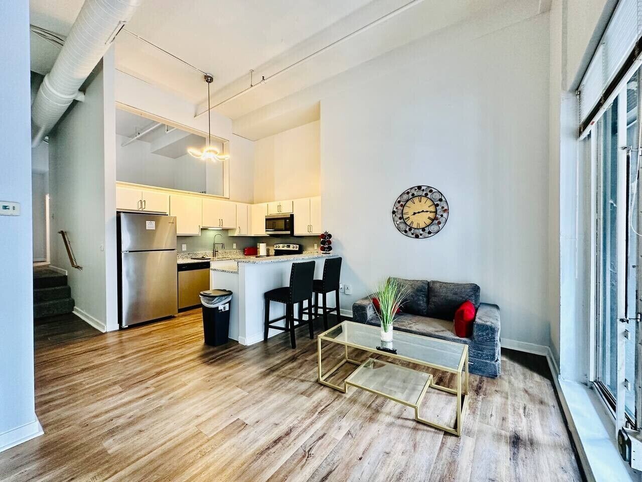 A modern kitchen with stainless steel appliances, a dining area with a glass coffee table, and a living room with a sofa and a decorative wall clock.