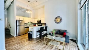 Una cocina moderna con electrodomésticos de acero inoxidable, un área de comedor con una mesa de centro de vidrio y una sala de estar con un sofá y un reloj decorativo en la pared.
