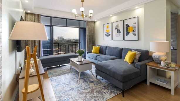 Un salon moderne avec un canapé gris, des coussins jaunes, une table basse et une vue sur la ville grâce à de grandes fenêtres.