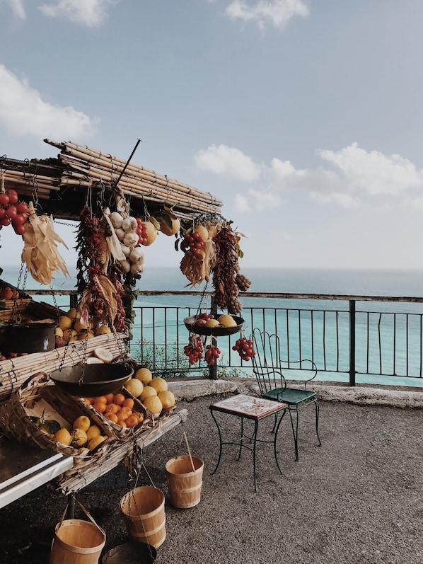 Un étal de marché situé en bord de mer, où l’on trouve divers fruits et légumes, une chaise, et d’où l’on peut admirer l’océan.