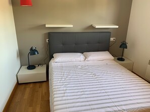 Ein ordentlich bezogenes Bett mit gestreifter Bettwäsche, zwei Nachttische mit Lampen und ein graues gepolstertes Kopfteil.