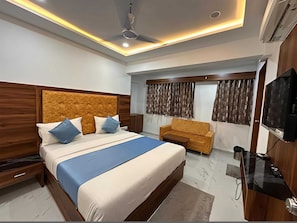 A modern hotel room with a large bed, a sofa, a TV, and a ceiling fan. - Deluxe Aura Inn (Ahmedabad)