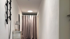 Un pasillo con suelo oscuro, pared de color claro, puerta blanca, alfombra beige, una mesita con lámpara y una cortina.