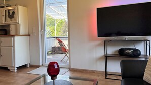 Una sala de estar con un televisor montado en la pared, una mesa de centro de vidrio con una copa de vino, y vista al exterior a través de un ventanal.