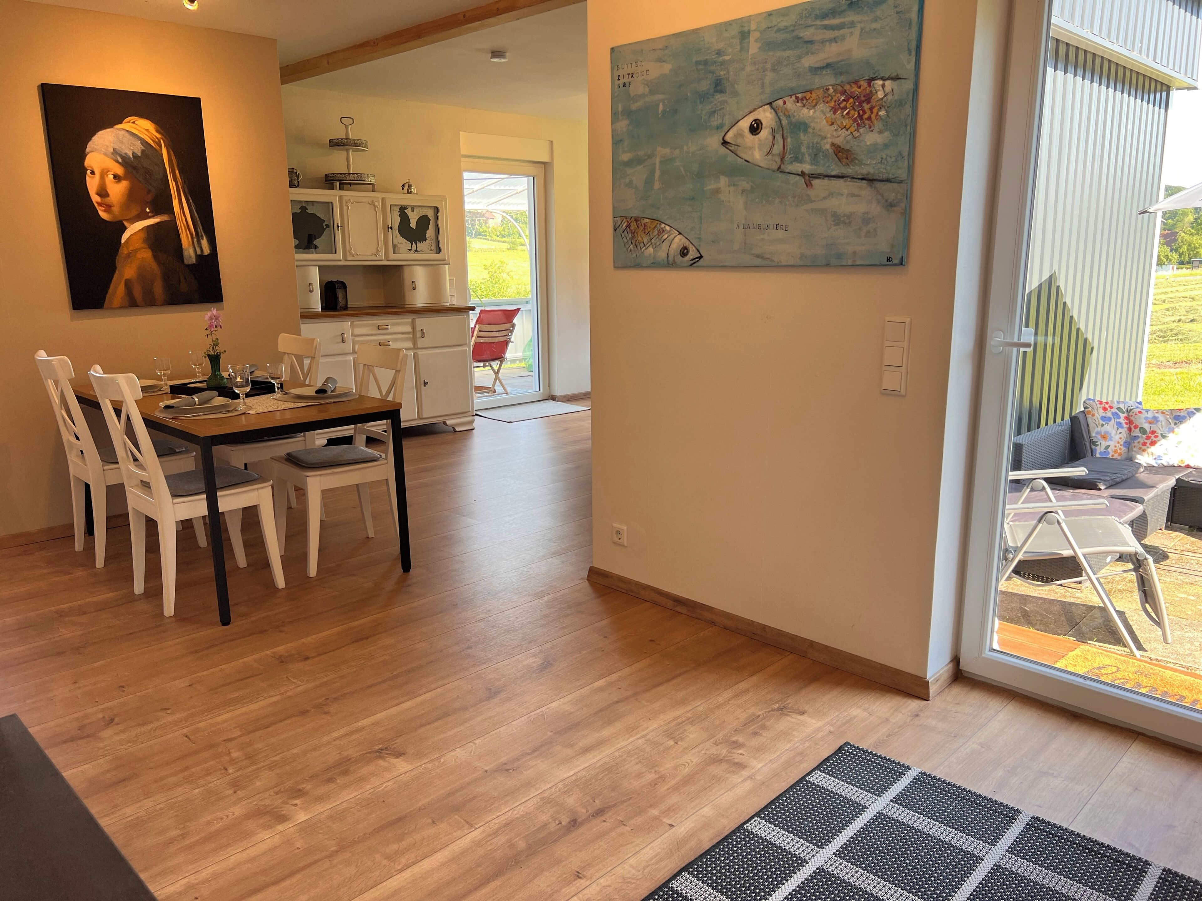 A dining area with a table set for four, a kitchen in the background, and a large painting on the wall.