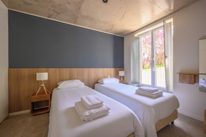 Une chambre d’hôtel avec deux lits, une tête de lit en bois, une table de chevet avec une lampe et une fenêtre donnant sur des arbres.