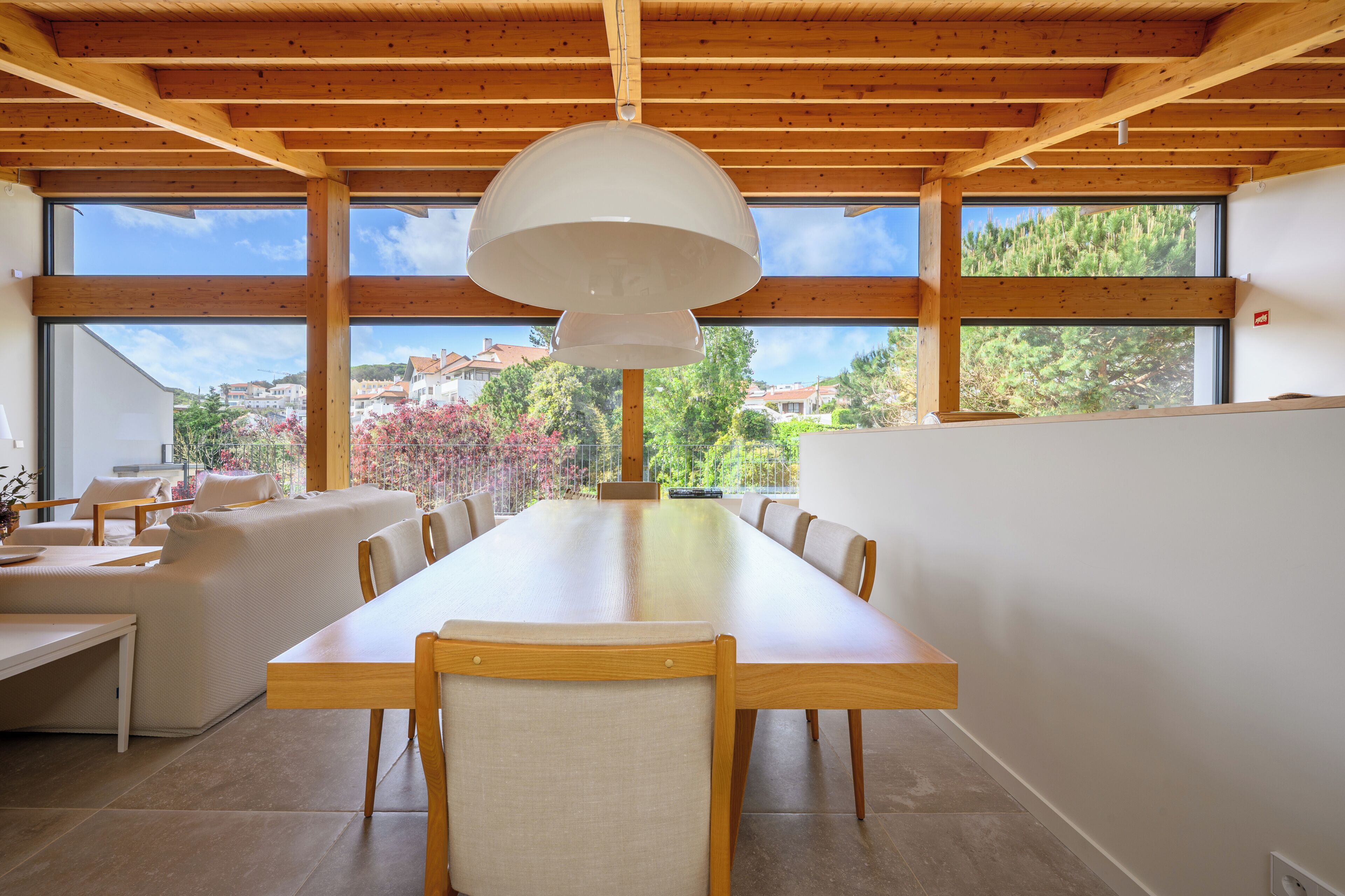 Een moderne eetruimte met een lange houten tafel, stoelen en een groot raam met uitzicht op huizen en bomen.