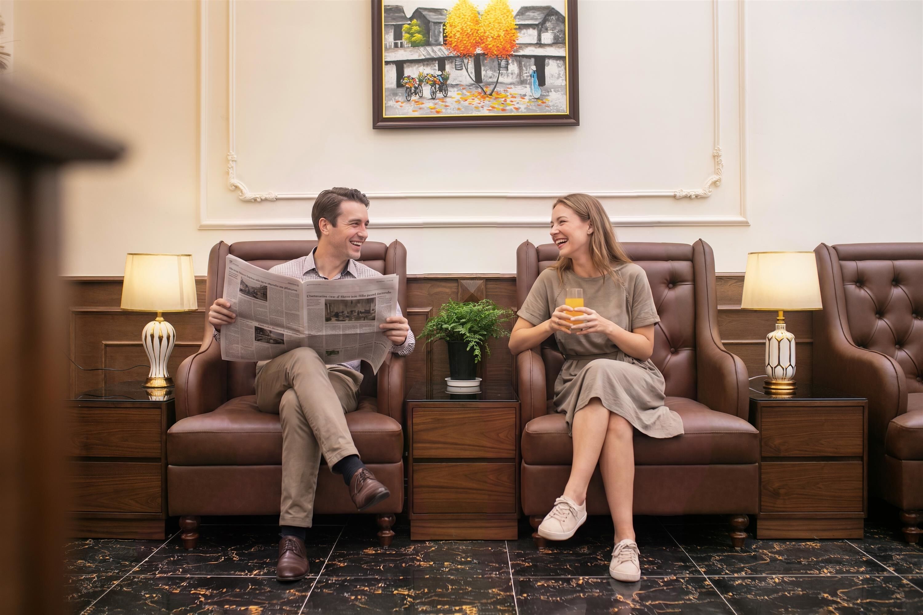 Two people seated on leather sofas in a room with a framed painting on the wall, a small potted plant, and a wooden side table with a lamp.