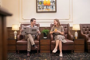 Two people seated on leather sofas in a room with a framed painting on the wall, a small potted plant, and a wooden side table with a lamp. - Bastra Classy Hotel (Hanoi)