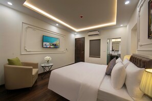 Habitación de hotel con cama, televisor montado en la pared, una pequeña mesa redonda con un jarrón de flores y una silla.