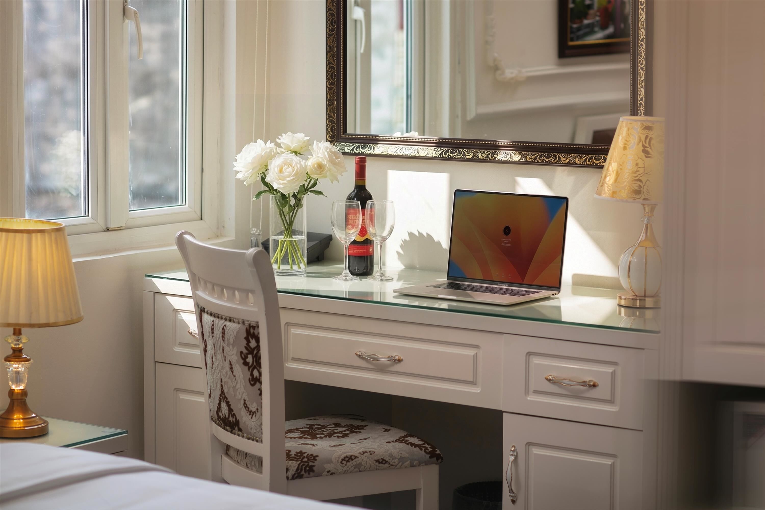 A room with a glass desk, a laptop, a vase with flowers, a bottle, and a lamp.